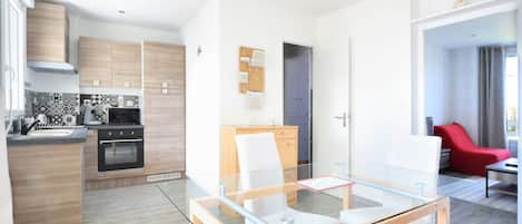 Una cocina moderna con armarios de madera, horno empotrado y microondas. Hay una mesa de comedor de vidrio con sillas blancas. La habitación tiene una silla roja y una mesita con camino de mesa rojo.
