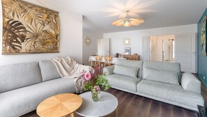 Een woonkamer met een grijze bank, een ronde houten salontafel en een bloemenvaas. Er is een plafondventilator en een wandlamp.