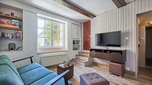 A modern living room with a sofa, ottoman, and a TV on a wooden cabinet. There is a window with a view of greenery, a radiator, and a staircase leading to another room. - Augustine Beach - Maison à 700 m de la plage (Courseulles-sur-Mer)