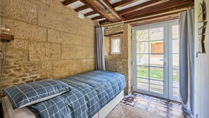 A bedroom with a bed, stone walls, wooden beams, a window, and a door leading to an outdoor area. - Augustine Beach - Maison à 700 m de la plage (Courseulles-sur-Mer)