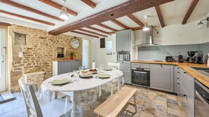 A kitchen with a stone wall, wooden beams, a round table with chairs, and modern appliances. - Augustine Beach - Maison à 700 m de la plage (Courseulles-sur-Mer)
