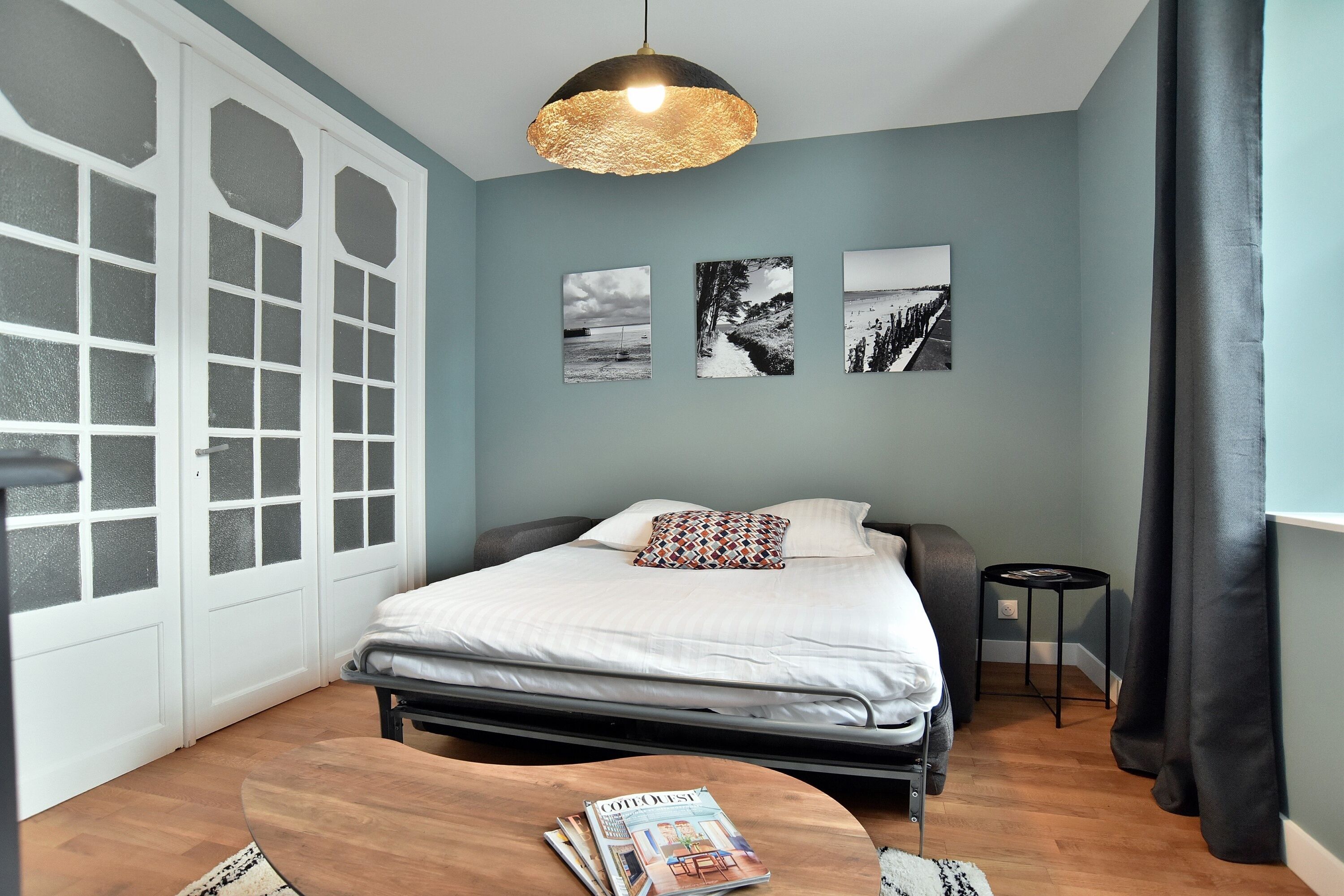 Ein Schlafzimmer mit einem Bett, einem runden Holztisch mit Zeitschriften, einem kleinen Beistelltisch und drei schwarz-weißen Fotografien an der Wand.