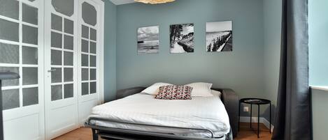 Ein Schlafzimmer mit einem Bett, einem runden Holztisch mit Zeitschriften, einem kleinen Beistelltisch und drei schwarz-weißen Fotografien an der Wand.