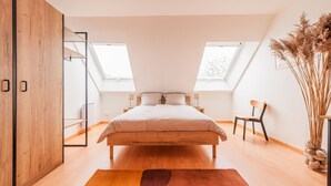 Un dormitorio con cama, tragaluces, un armario de madera, una silla y un jarrón con plantas secas.