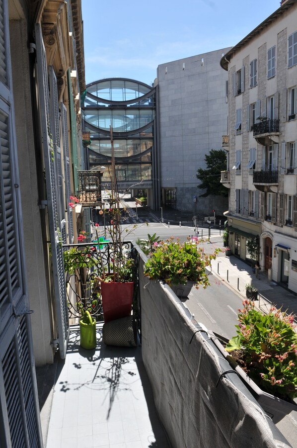 A narrow urban street with buildings, a glass structure, and potted plants on a balcony.