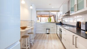A modern kitchen with a large window, white cabinets, and a black countertop. There are two stools and a trash can. - Great private terrace in the heart of the city centre  (Málaga)