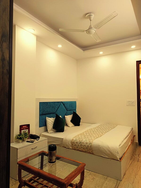 A bedroom with a bed, a bedside table with a phone and a remote, a ceiling fan, and a nightstand with a plant.