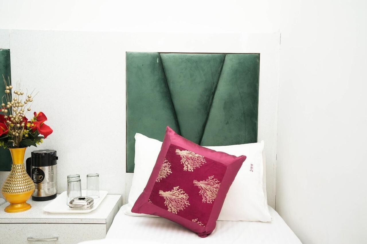 A neatly arranged hotel room with a green upholstered headboard, a white pillow with a red and gold pattern, a white bedside table with a vase of flowers, a black and silver electric kettle, and two glass containers.