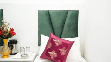 A neatly arranged hotel room with a green upholstered headboard, a white pillow with a red and gold pattern, a white bedside table with a vase of flowers, a black and silver electric kettle, and two glass containers.