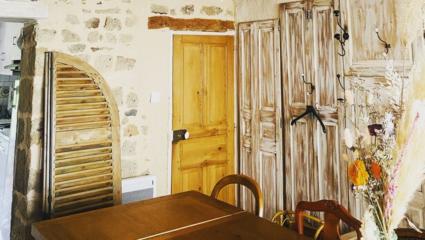 A rustic dining area with a wooden table, a ceiling fan, and a door with a wooden shutter. - Maison de Caractère à Saint-nectaire (Saint-Nectaire)