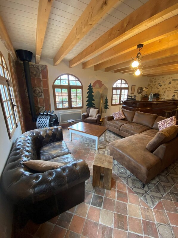 Un salon chaleureux avec un canapé en cuir, une table basse en bois et un divan marron. De grandes fenêtres à arc, une cheminée et un mur de pierre orné d’une fresque représentant une montagne complètent l’ensemble.