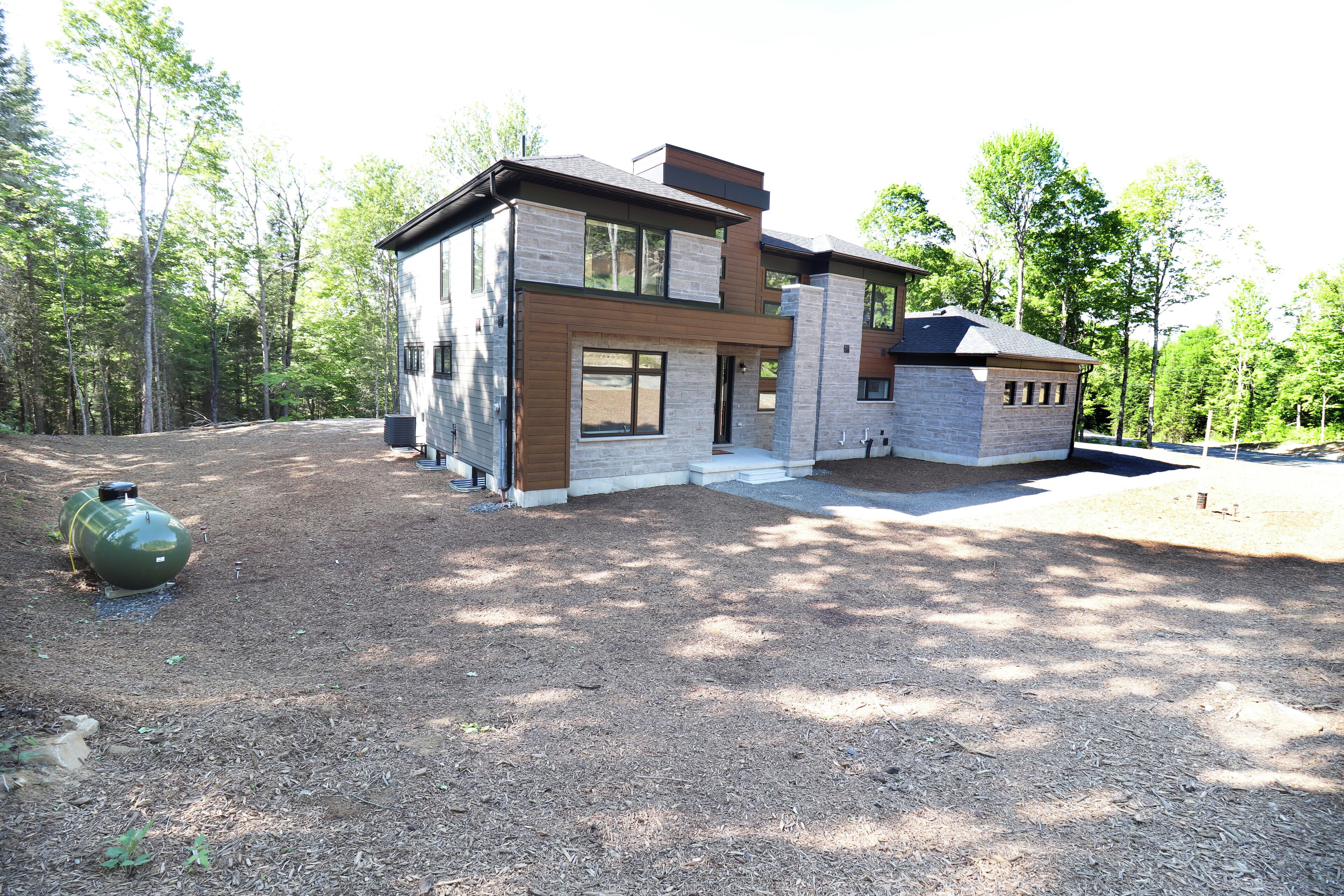 Une maison moderne à deux étages, dotée d’une façade en pierre et de grandes fenêtres, et entourée d’une zone boisée.