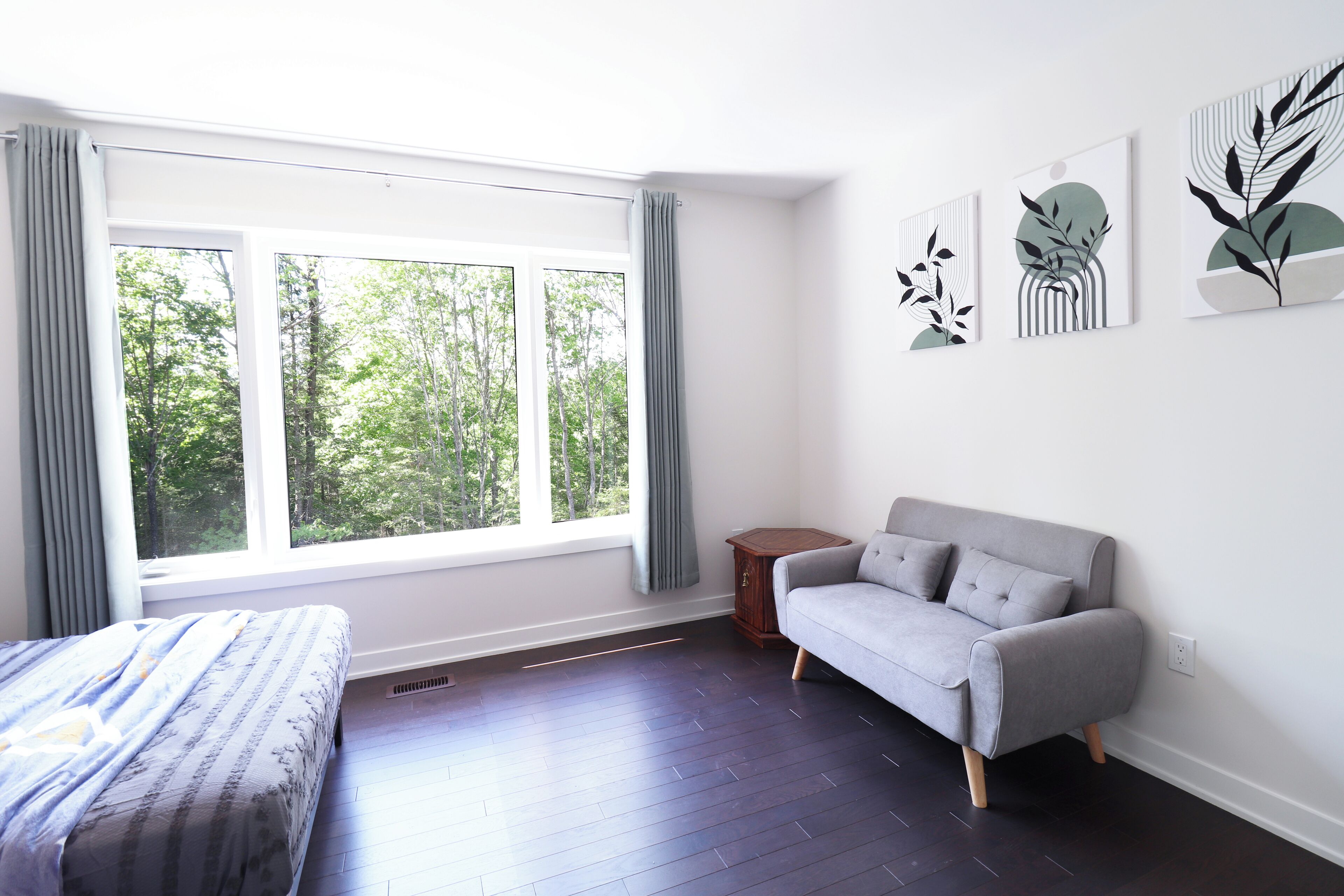 Une chambre moderne avec un lit, un canapé gris, une fenêtre avec des rideaux et trois œuvres d’art accrochées au mur.