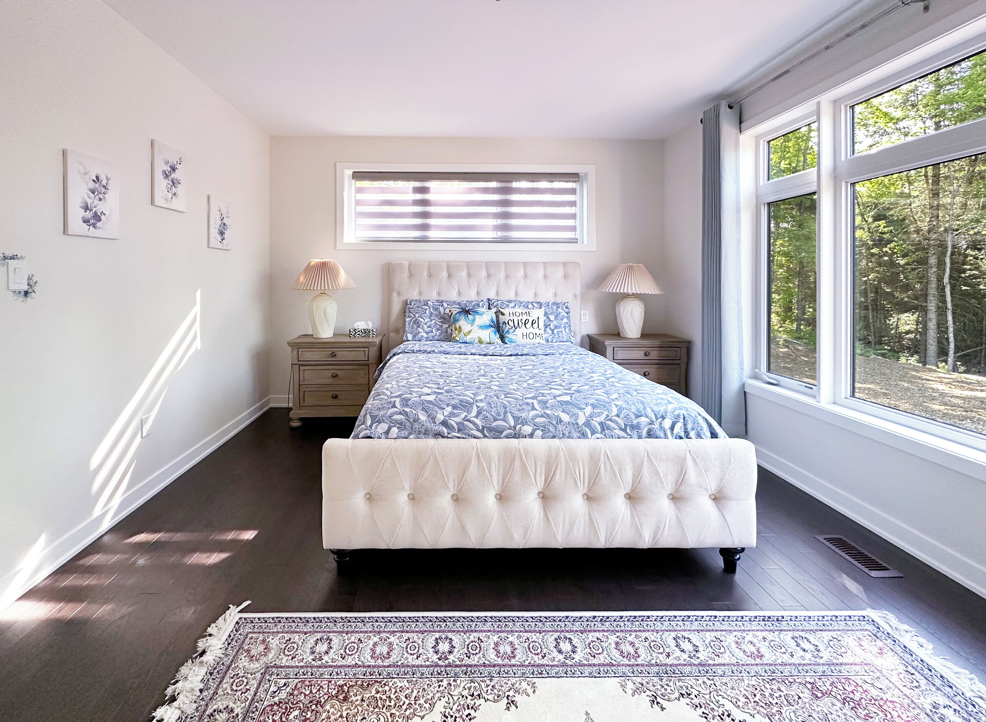 A bedroom with a large bed, two nightstands, a window with blinds, and a view of trees.