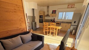 A modern living room with a sofa, wooden coffee table, and a TV. A kitchen with wooden cabinets and a dining area is visible in the background. - New single-storey chalet in Gérardmer (Xonrupt-Longemer)