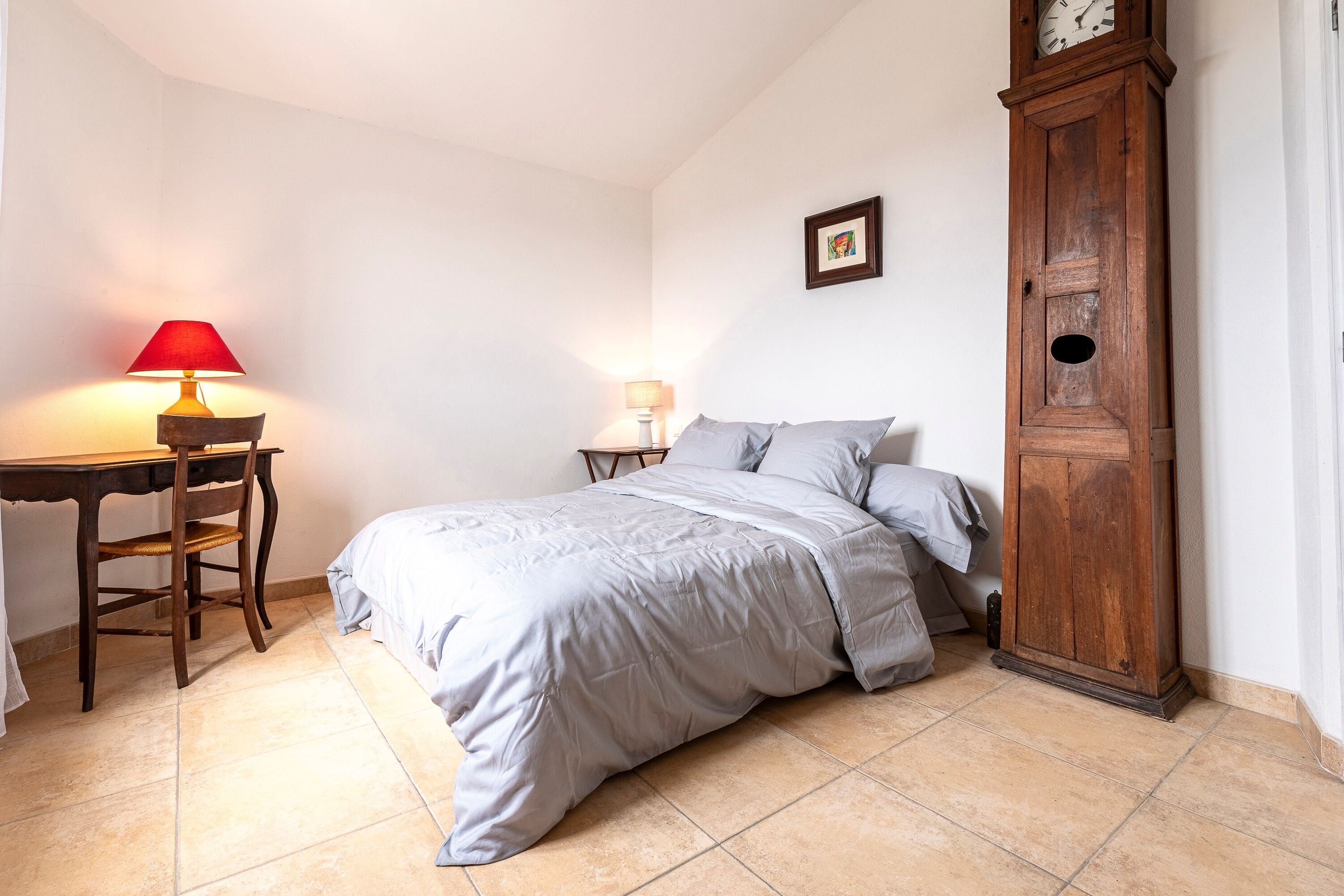 A bedroom with a bed, a desk with a lamp, a wooden cabinet, and a wall clock.