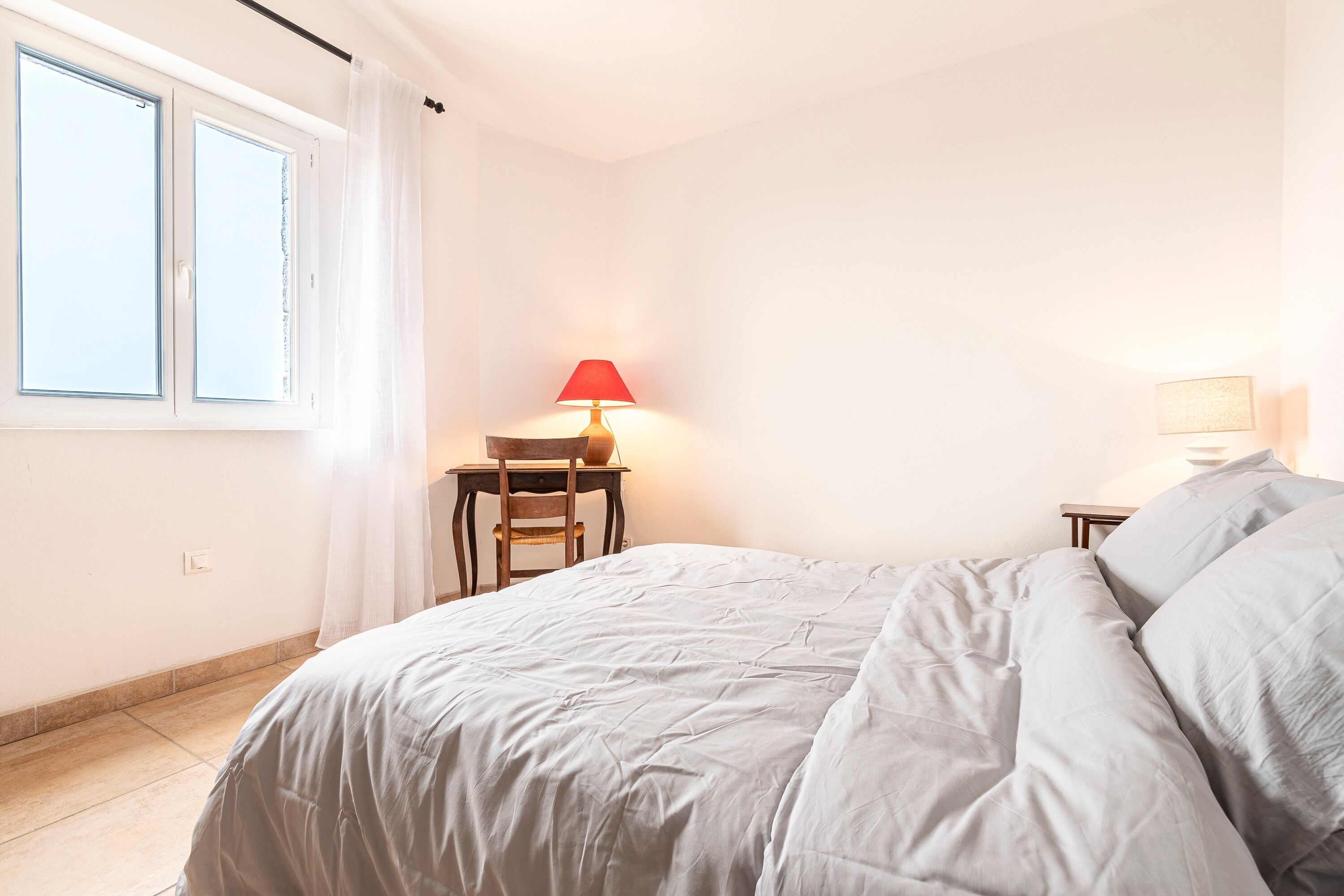 Um quarto com cama, uma cadeira, uma mesa com abajur e uma janela com cortinas.