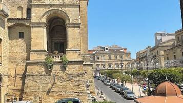 A historic stone building with a bell tower, a street with parked cars, and modern buildings in the background.