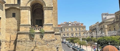 A historic stone building with a bell tower, a street with parked cars, and modern buildings in the background.