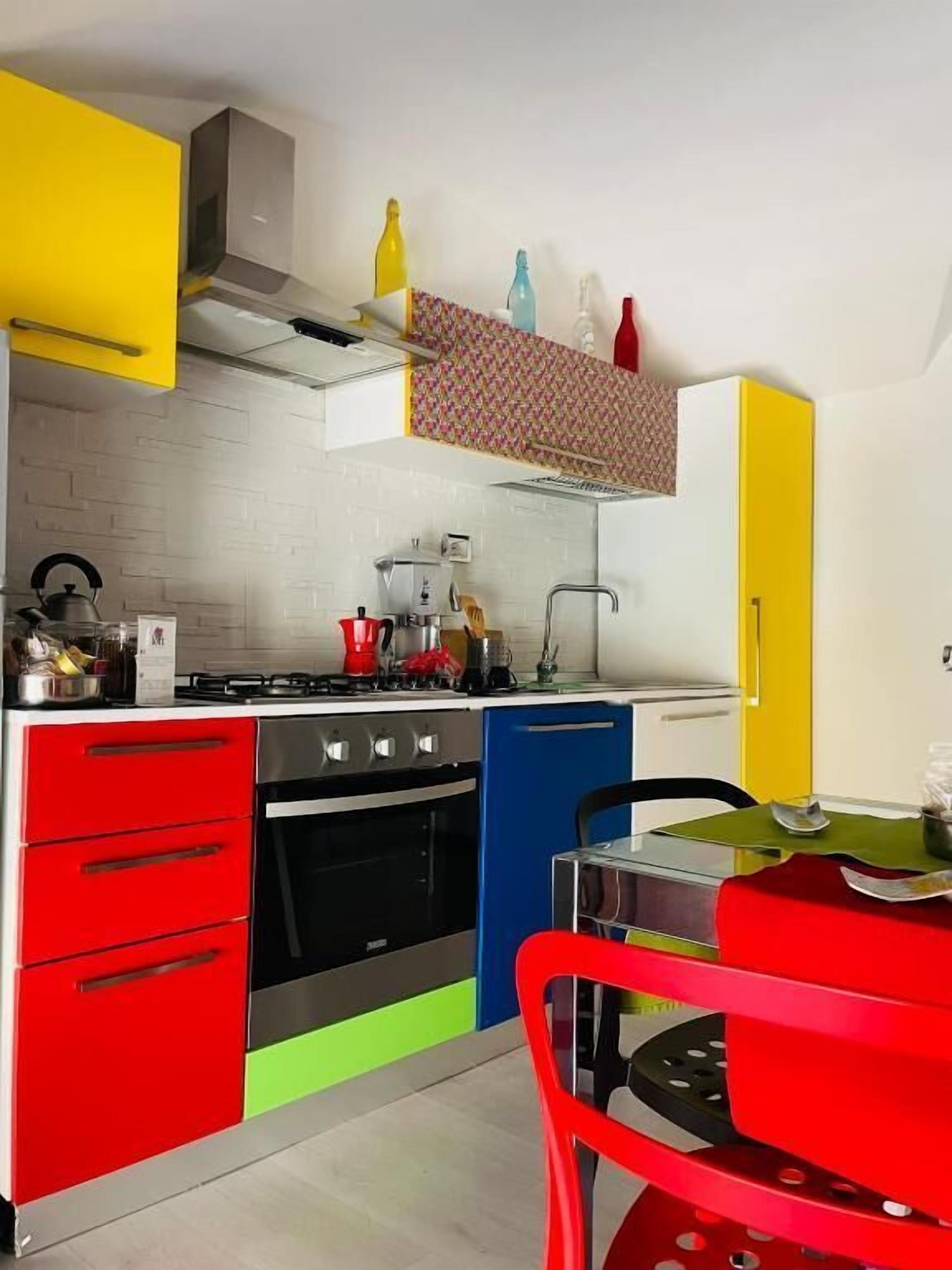 Een compacte keuken met een gasfornuis, oven en moderne apparaten. Er is een eethoek met een rode stoel en een tafel.