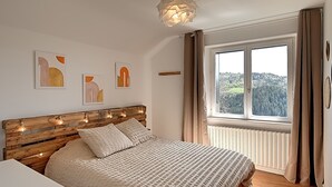 Ein Schlafzimmer mit einem Holzkopfteil, einem großen Bett mit gemustertem Bettbezug, einem Nachttisch und einem Fenster mit Blick auf ein bewaldetes Gebiet.
