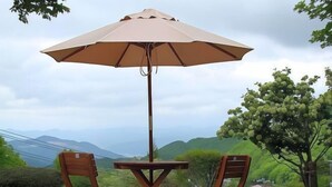 Una mesa y dos sillas bajo un paraguas grande, con colinas verdes y un cielo nublado de fondo.