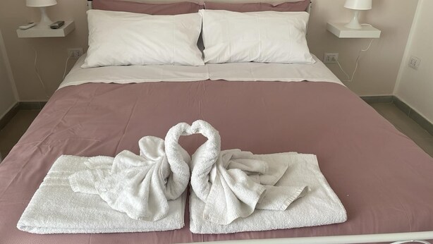 A neatly made bed with a pink bedspread and white pillows. There are two white lamps on either side of the bed. - Una Notte a Napoli (Naples)