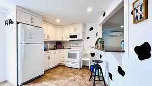 Une cuisine moderne avec des meubles blancs, un réfrigérateur, un four à micro-ondes et une cuisinière. On peut voir un petit tabouret et une bouteille d’eau sur le comptoir.