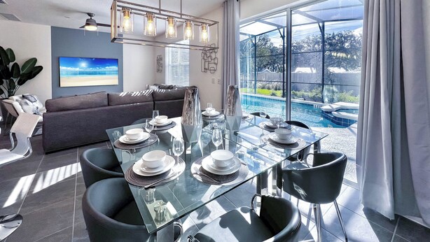 A modern dining area with a glass table set for six, a gray sofa, and a large window overlooking a pool. - BV310MB - Disney & Universal heroes - 16 guests (Kissimmee)