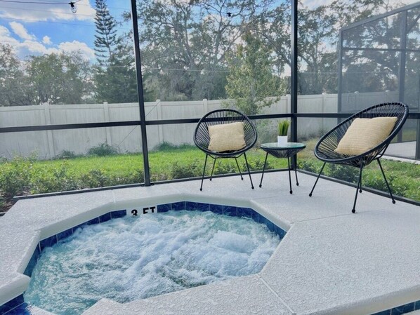 Ein Außenbereich mit Whirlpool, zwei Korbstühlen und einem kleinen Tisch, umgeben von einer Glaseinfriedung mit Blick auf einen Holenzaun und Grünpflanzen.