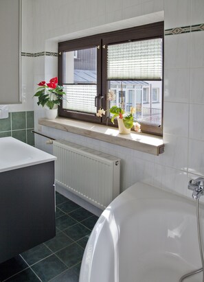 Un baño con bañera, ventana con persianas, radiador y planta en maceta.