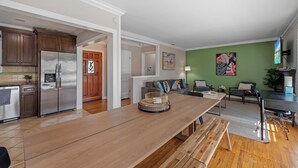 A modern kitchen with stainless steel appliances, a wooden dining table, and a living area with a green wall and artwork. - Ladoga's Hideway, Gorgeous Pool, Game Room, Fire Pit, Private Backyard,BBQ Grill (Long Beach)