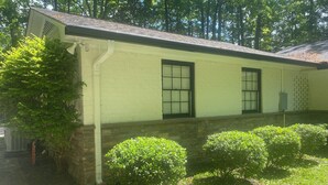 A single-story building with a light-colored exterior, a brick foundation, and a sloped roof. There are three windows with dark frames, and a small porch area with a white railing. - Apartment in Downtown Clemson. Just a few minutes to Campus & Football Stadium. (Clemson)