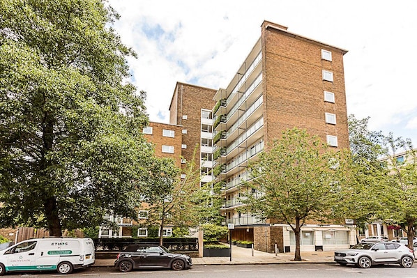 A multi-story residential building with a brick facade, balconies, and a parking area with cars.