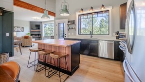 A modern kitchen with a central island, bar stools, and a large window providing a view of the outdoors. - Glacier Natl Park & River Views, Dog Friendly (Hungry Horse)