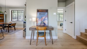 A dining area with a wooden table and chairs, a painting of Montana's Midnight Serenade, and a view of the outdoors through large windows. - Glacier Natl Park & River Views, Dog Friendly (Hungry Horse)