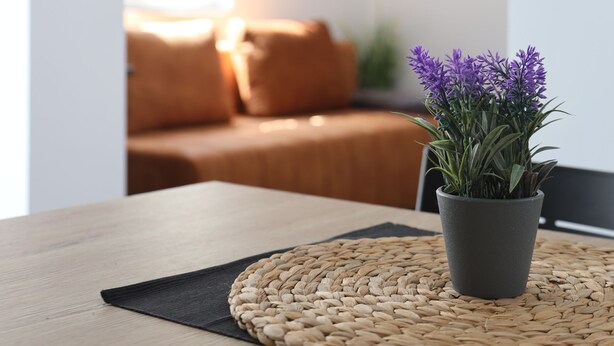 Una mesa de madera con un posavasos tejido y una maceta con flores púrpuras.
