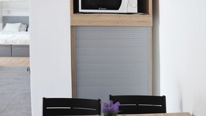 Un comedor con una mesa de madera, sillas negras, un mantel tejido y una planta en maceta. Se observan un microondas y una tetera en un estante superior.