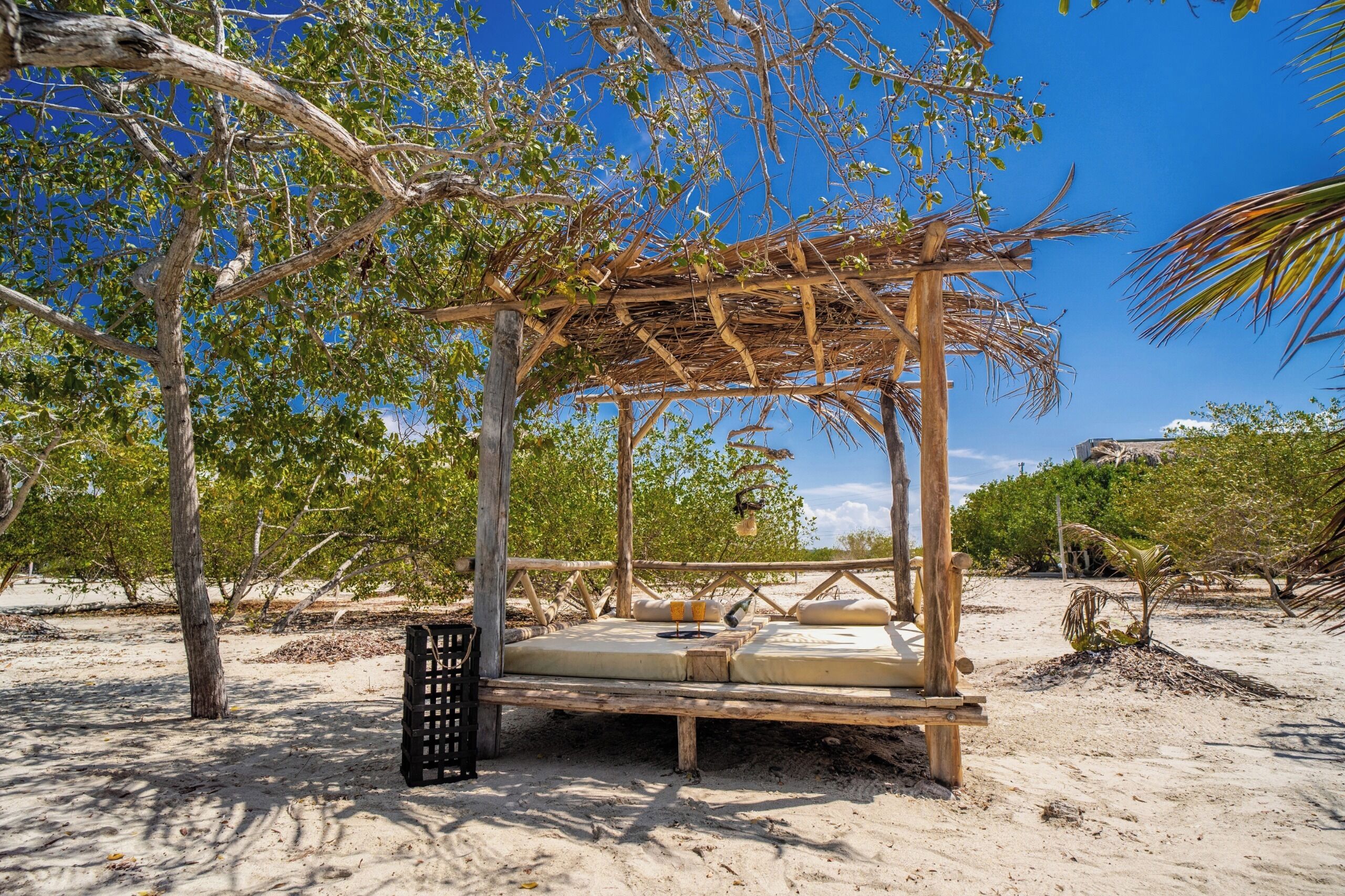 A wooden gazebo with a bed and a small table, surrounded by trees and sand.