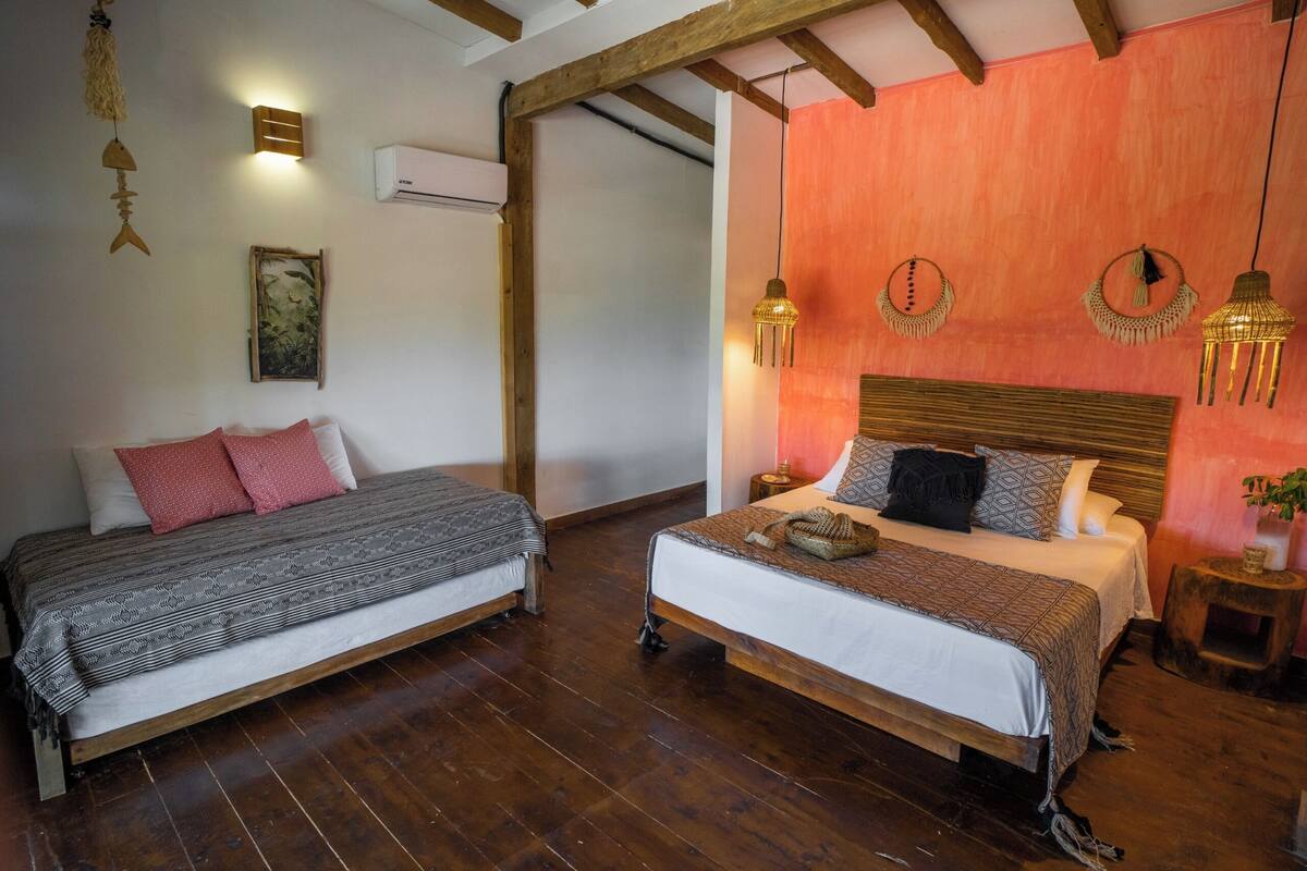 A bedroom with two beds, wooden beams, a red accent wall, and hanging light fixtures.