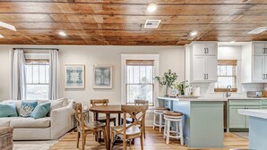 Una sala de estar acogedora con techo de madera, un comedor con mesa y sillas de madera, y una cocina con armarios blancos y una isla azul.