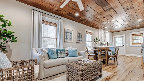 Una sala moderna con techo de madera, un sofá color beige con cojines azules, una mesa de centro de mimbre y un comedor con sillas y mesa de madera.
