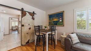 A dining area with a wooden table and chairs, a sliding barn door, a painting of a sunset, a leather sofa, and a window with blinds.