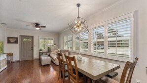A dining area with a wooden table, chairs, and a pendant light. There is a ceiling fan and a window with blinds.