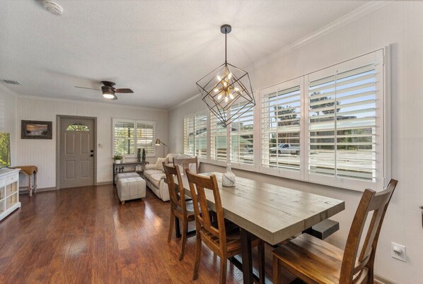 A dining area with a wooden table, chairs, and a pendant light. There is a ceiling fan and a window with blinds.