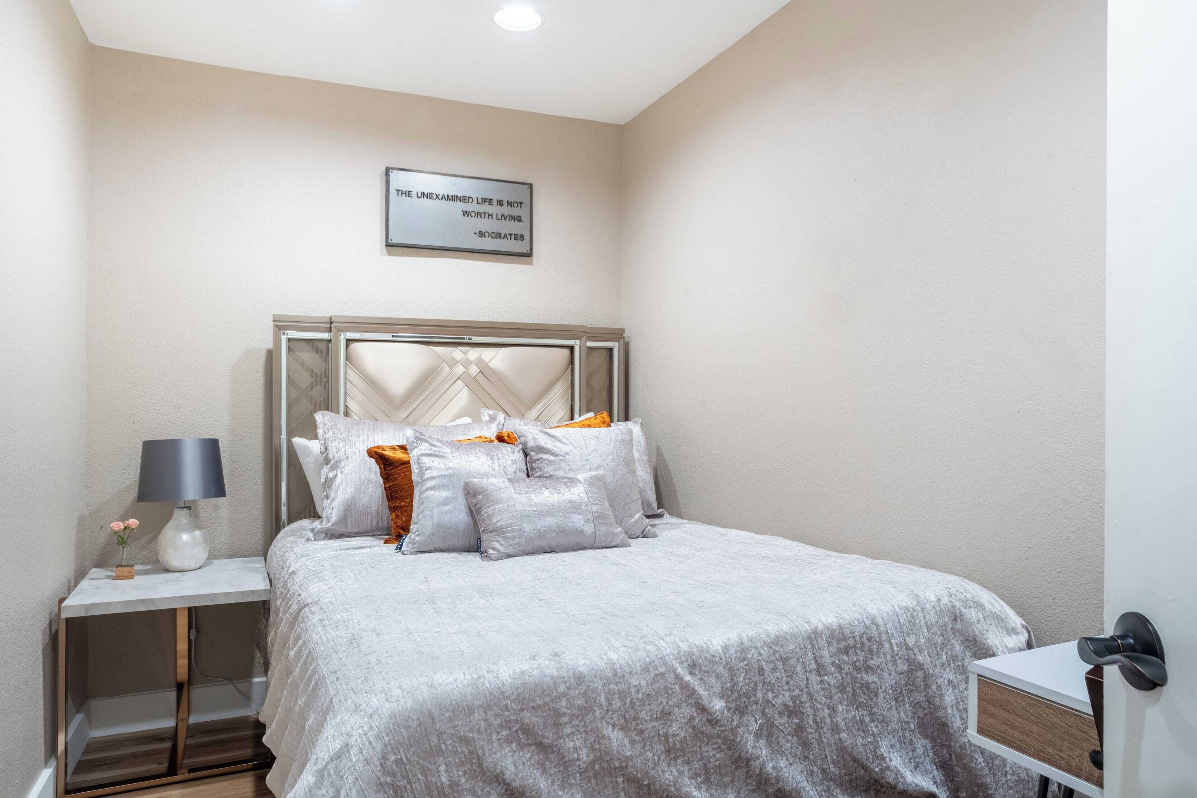 A neatly arranged bedroom with a bed, bedside table, lamp, and a motivational quote on the wall.