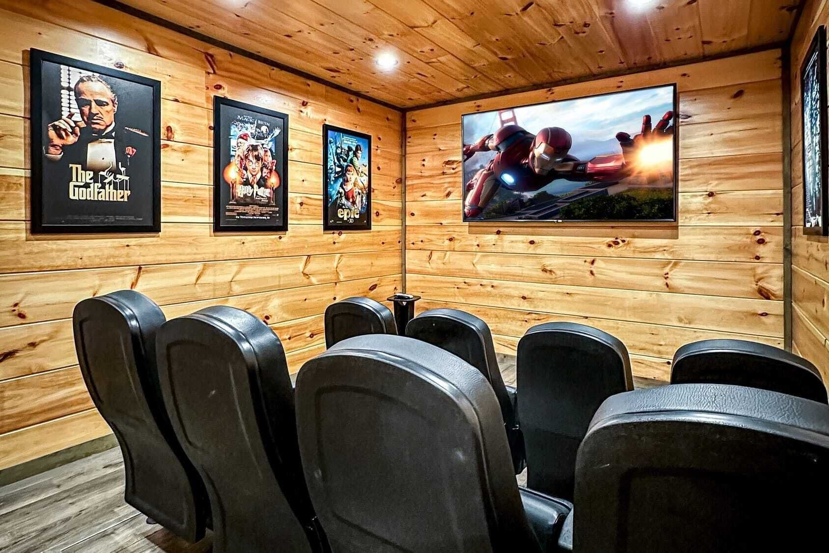 A wooden-paneled room with black cinema-style seating and three movie posters on the wall.