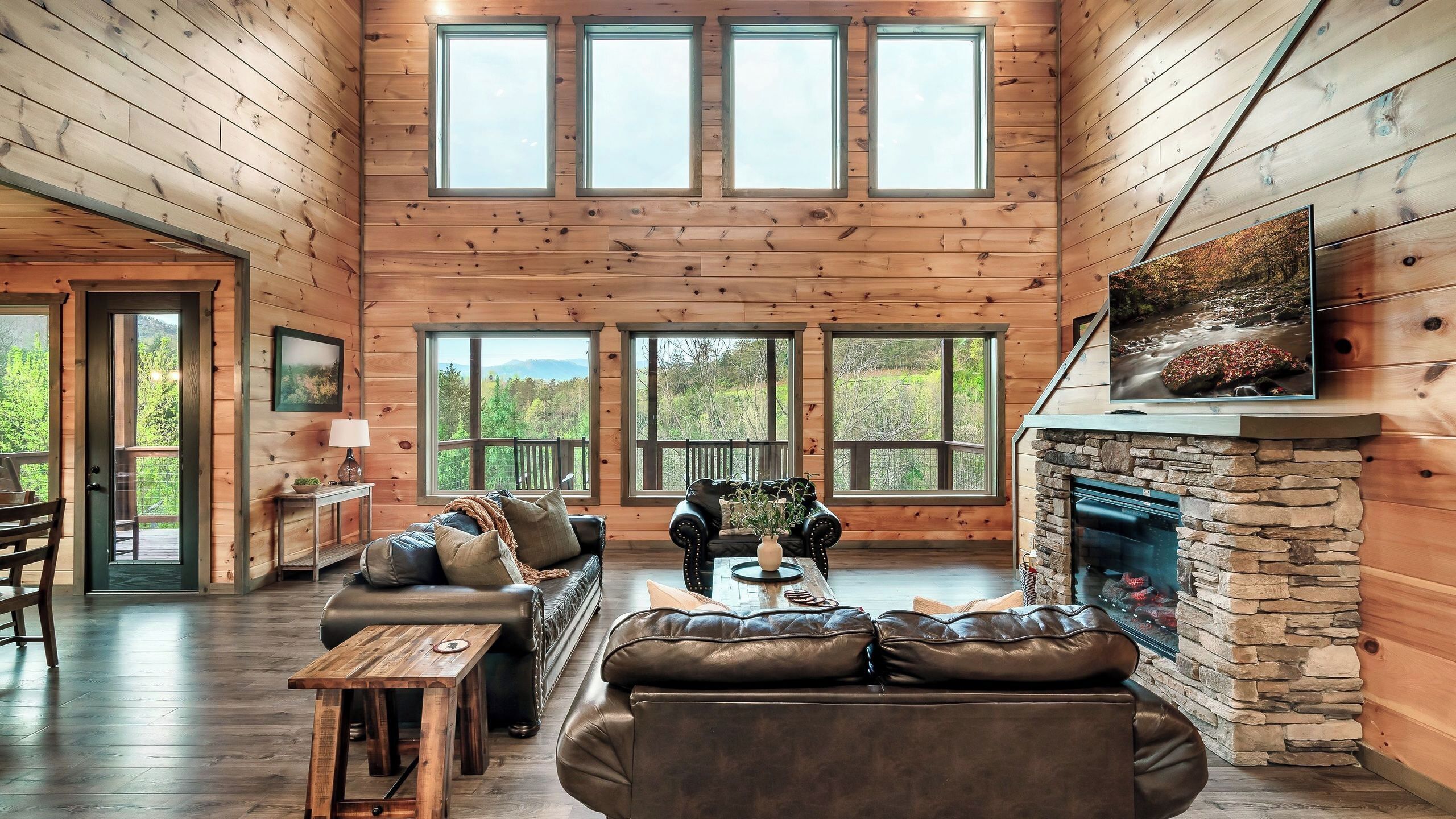 A spacious log cabin interior with a stone fireplace, leather sofas, and large windows offering scenic views.