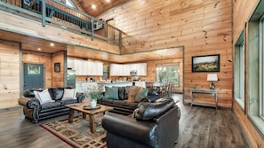 A spacious living area with a high ceiling, wooden walls, a dining area, and a kitchen with white cabinets. - Smoky Horizon by Avantstay Game Room, Movie Theater, Deck w Hot Tub Views! (Sevierville)