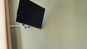 A room with a desk, a television mounted on the wall, a trash bin, and a wicker ottoman.